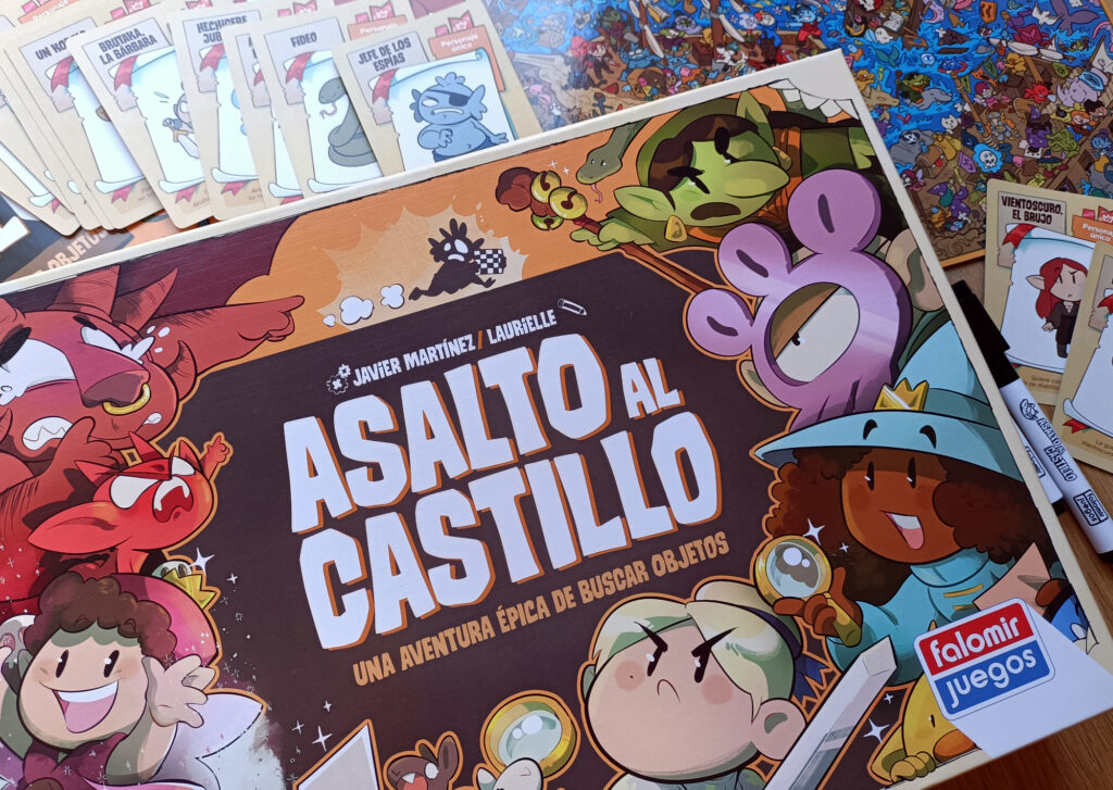 Foto de la caja del juego Asalto al Castillo con un tablero, cartas y rotuladores de fondo. El subtítulo del juego es Una aventura épica de buscar objetos