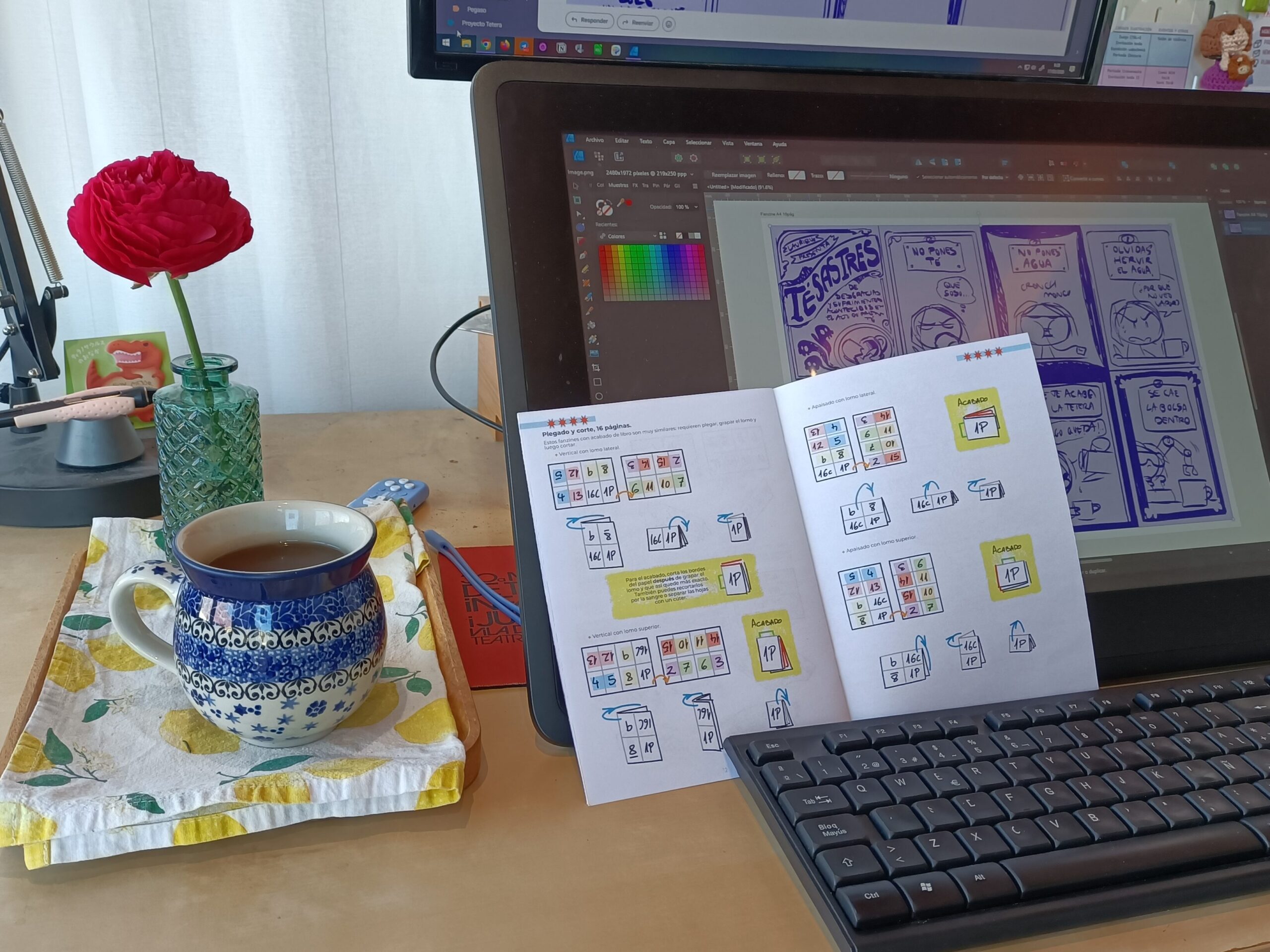 Foto de mi mesa de trabajo con un té, una flor muy aesthetic, mi tableta de trabajo con el dibujo abierto y un fanzine con instrucciones para plegar fanzines abierto sobre la tablet