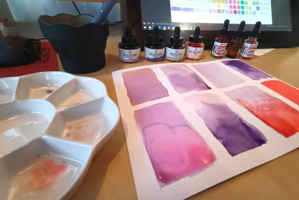 Foto de la misma paleta de la imagen anterior, botes de anilinas, una tablet con las muestras escaneadas y varios swatches de acuarela morada, violeta y roja en un papel