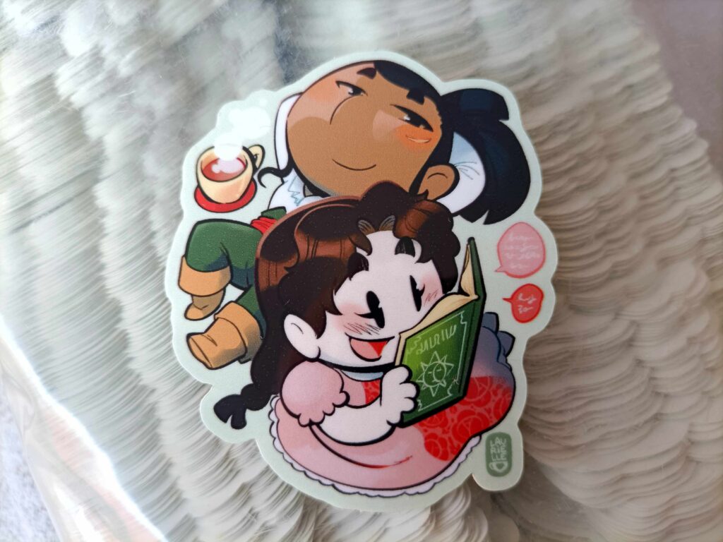 Foto de una pegatina de Cam tirada en el suelo con cara de satisfacción mientras mira a Margarita, que está leyendo un libro muy contenta. La pegatina está sobre un montón de pegatinas más.